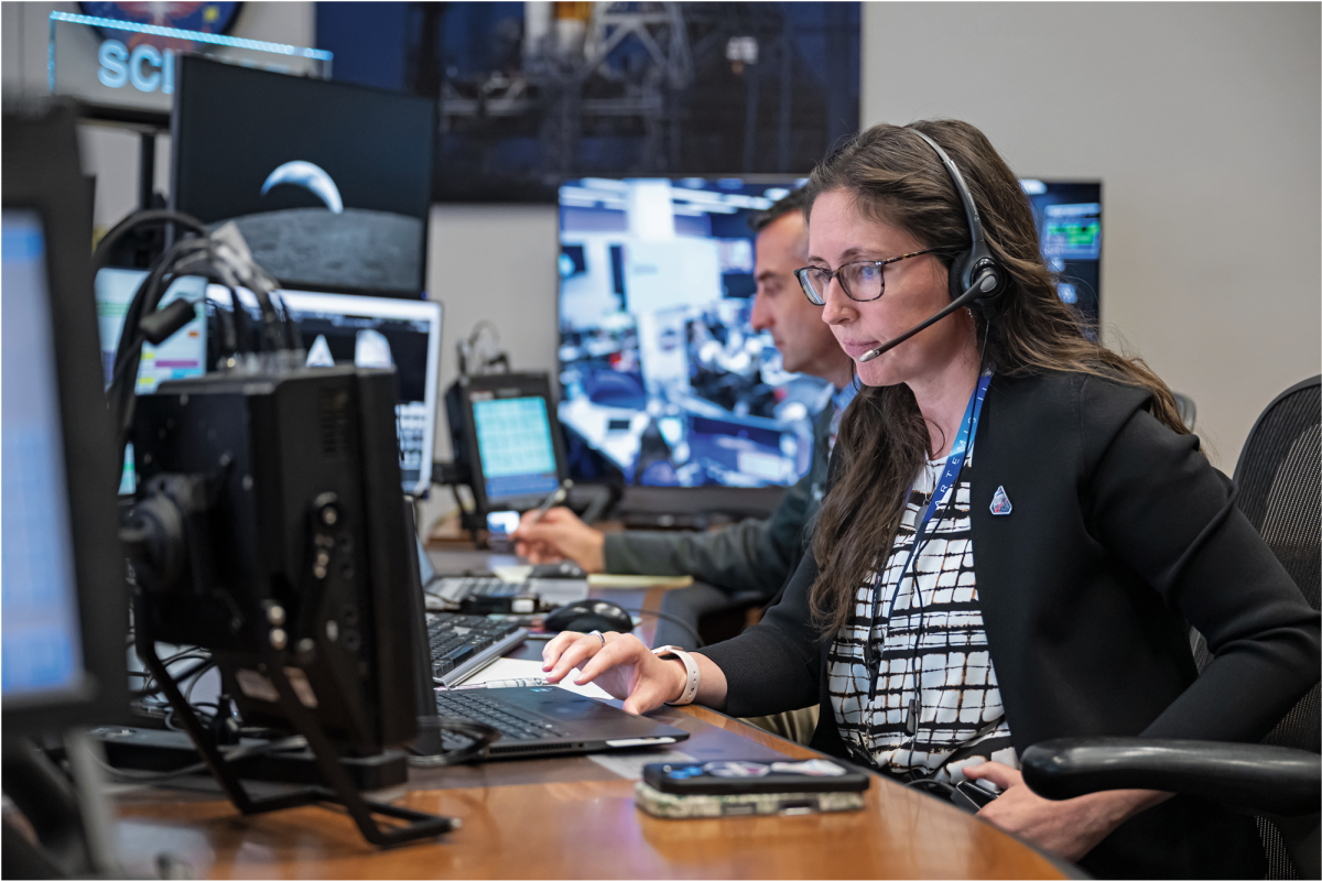 A woman sits at a desk working on a computer with multiple screens